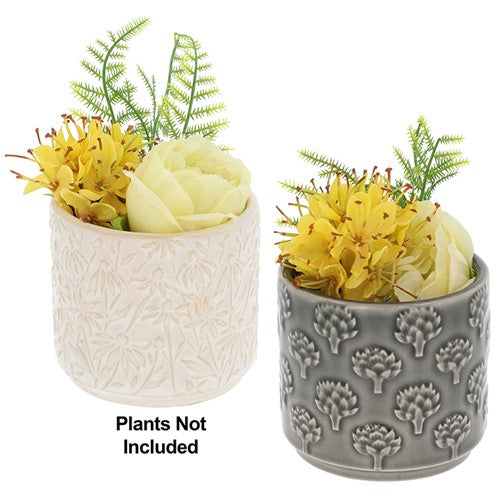 Two cylindrical ceramic planters shown side by side on a white background, each filled with decorative faux yellow flowers and green ferns. The left planter is off-white with an embossed floral pattern, and the right planter is gray with a matching embossed tree-like pattern.