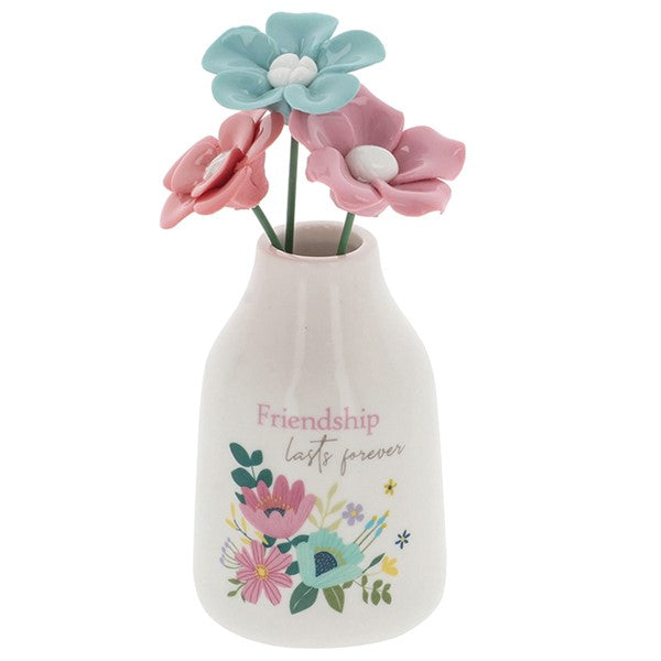 A small white ceramic vase with the words 'Friendship lasts forever' and a colorful floral illustration, holding pink and light blue decorative flowers on a white background.