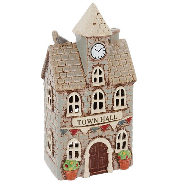 A small decorative ceramic model of a town hall building with a clock face on the tower, arched windows, a brown door, potted plants at the base, and a white background.