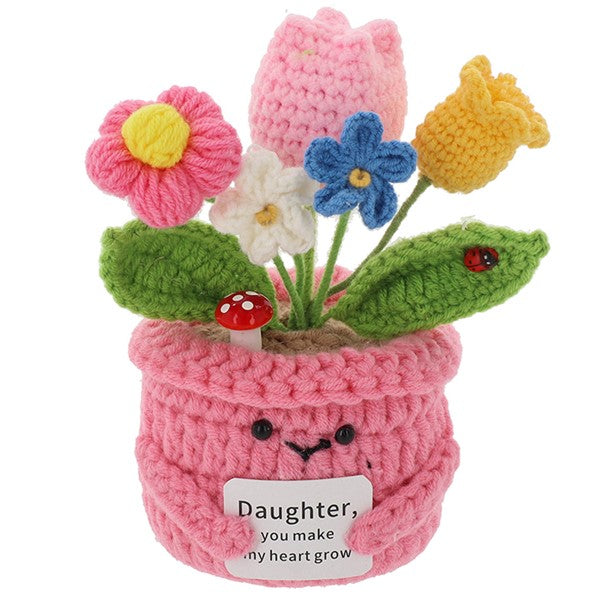 A crocheted flower pot with handmade yarn flowers in pink, blue, yellow, white, and green leaves, featuring a cute embroidered face and a message card that reads 'Daughter, you make my heart grow'.