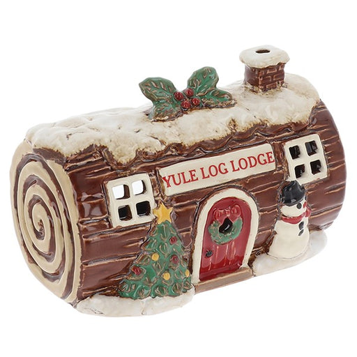 A ceramic Christmas ornament shaped like a log cabin labeled 'YULE LOG LODGE,' with faux snow on top, a red door, green wreath and holly, a small tree, and a snowman figure on a white background.