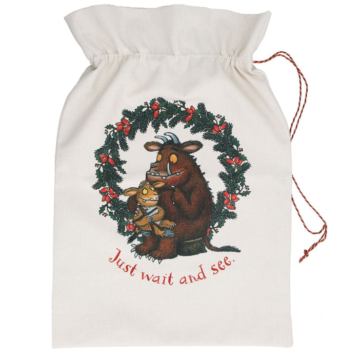 An off-white canvas Christmas stocking bag with a drawstring closure, featuring a printed illustration of a brown grumpy cow and a small yellow bird inside a green holly wreath with red berries. The text 'Just wait and see.