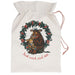 An off-white canvas Christmas stocking bag with a drawstring closure, featuring a printed illustration of a brown grumpy cow and a small yellow bird inside a green holly wreath with red berries. The text 'Just wait and see.