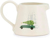An off-white ceramic milk pitcher featuring a whimsical illustration of a green vintage car carrying a green Christmas tree on its roof, with light gray pine trees in the background.