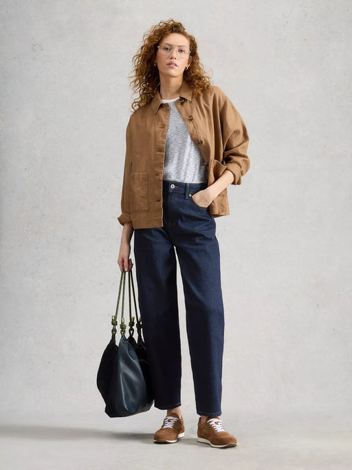 A woman stands against a light gray textured wall wearing a tan overshirt, white textured top, dark blue wide-leg jeans, and brown sneakers, while holding a large black tote bag.