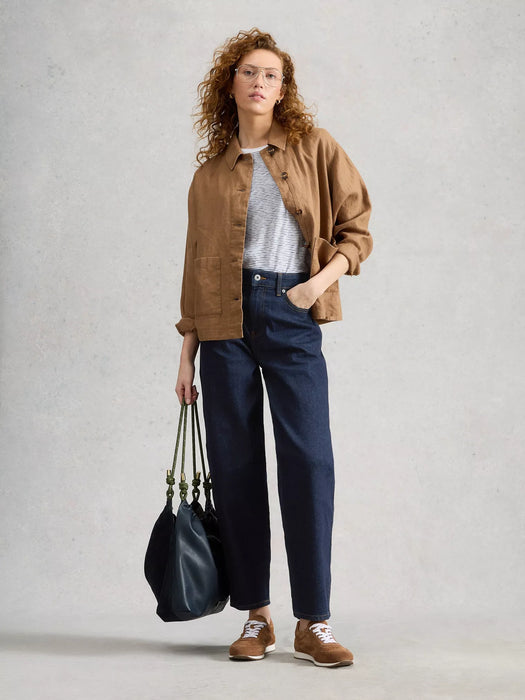 A woman stands against a light gray textured wall wearing a tan overshirt, white textured top, dark blue wide-leg jeans, and brown sneakers, while holding a large black tote bag.