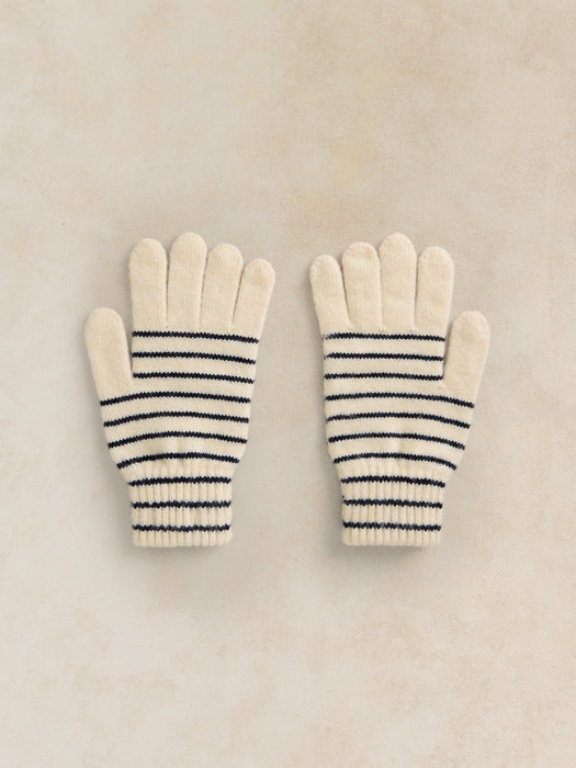 A pair of cream knit gloves with thin navy blue horizontal stripes, laid flat on a textured beige background.
