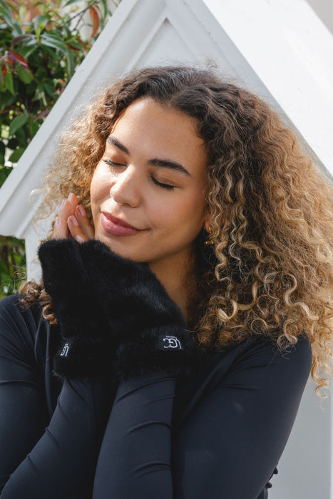 A woman with curly brown hair wearing a black long-sleeve top and a fluffy black faux-fur collar, posed in front of a white geometric background.