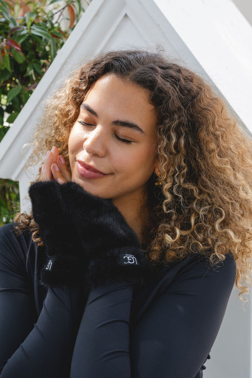 A woman with curly brown hair wearing a black long-sleeve top and a fluffy black faux-fur collar, posed in front of a white geometric background.