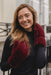 A smiling woman wearing a pair of deep burgundy faux fur gloves and a matching burgundy scarf, paired with a black long-sleeve top, set against a softly blurred urban background.
