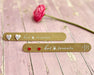 Two gold-toned rectangular tags with rounded ends on a distressed pink wooden surface. One tag has two silver glitter hearts and the other has two red hearts, both printed with the words 'hot tomato'.