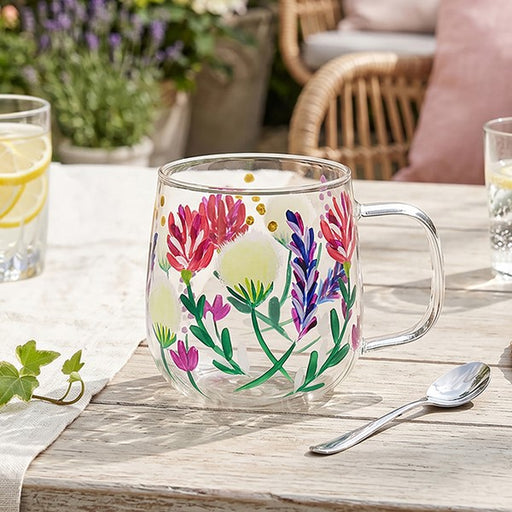 A clear glass mug with a floral hand-painted design featuring pink, purple, green, and yellow flowers, set on a rustic wooden table in an outdoor patio setting.