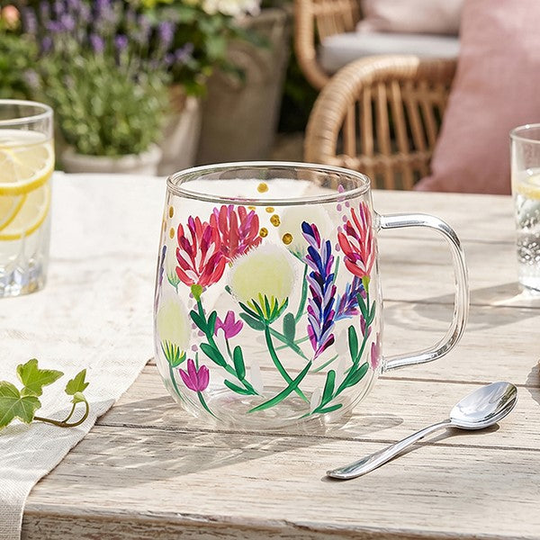 A clear glass mug with a floral hand-painted design featuring pink, purple, green, and yellow flowers, set on a rustic wooden table in an outdoor patio setting.