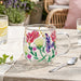 A clear glass mug with a floral hand-painted design featuring pink, purple, green, and yellow flowers, set on a rustic wooden table in an outdoor patio setting.