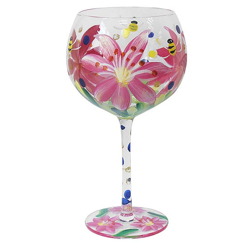 A clear glass wine glass decorated with a hand-painted pink lily flower, green leaves, blue dots, yellow and black bees, and a matching floral pattern on the base, shown against a plain white background.
