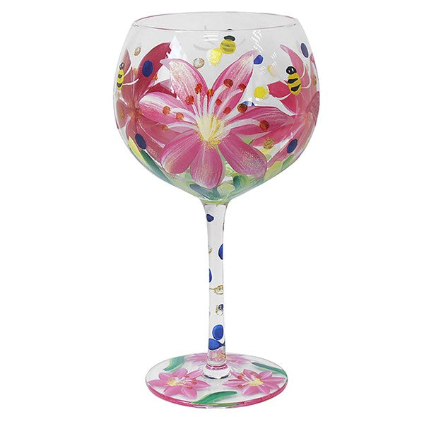 A clear glass wine glass decorated with a hand-painted pink lily flower, green leaves, blue dots, yellow and black bees, and a matching floral pattern on the base, shown against a plain white background.