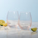 Two clear stemless wine glasses with a textured, ribbed pattern, displayed on a light marble surface with lime wedges and ice cubes against a soft light blue background.