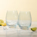 Two clear stemless wine glasses with a textured, ribbed pattern, displayed on a light marble surface with lime slices and ice cubes against a warm cream background.
