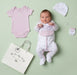 A baby laid on a pale green background with a light pink bodysuit, footed sleeper, hat, and glove arranged around them, plus a white shopping bag printed with the brand name 'Nooka Baby Boutique'. A real baby is present, so this is Lifestyle rather than Product/Packaging, even though the setup is styled like a product flat lay.