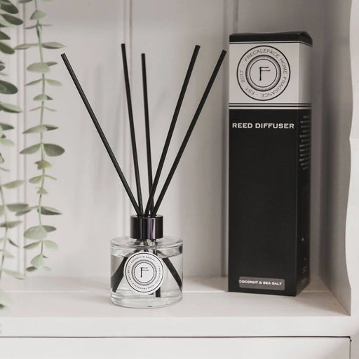 A clear glass reed diffuser bottle with black reeds and a black-and-white label sits on a white shelf beside its tall black product box. A hanging green plant is visible at the left edge.