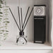 A clear glass reed diffuser bottle with black reeds and a black-and-white label sits on a white shelf beside its tall black product box. A hanging green plant is visible at the left edge.