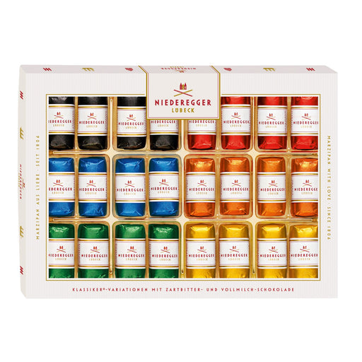 A boxed assortment of Niedergger chocolate tart and milk chocolate pieces in a white gift box with gold trim. The front shows multiple rows of small wrapped pieces in assorted colors including black, blue, green, orange, red, and yellow, with the brand name Niedergger visible on the packaging.